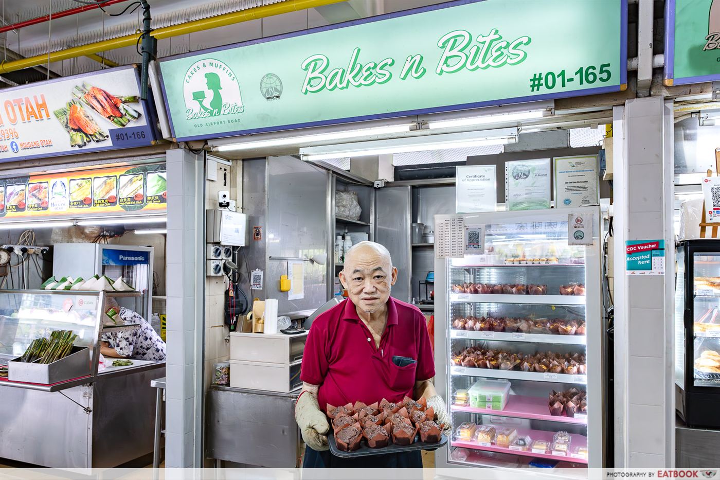 The Bakes n Bites storefront at Old Airport Road Food Centre, with owner Christopher and a chiller of freshly baked muffins